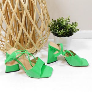 May include: A pair of bright green heeled sandals with a square toe and strappy design. The sandals have a textured, snake-like pattern and a chunky heel. The shoes are set against a white background with a decorative wicker basket and a small potted plant.