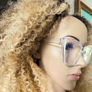 K&ouml;nnte beinhalten: Ein klarer Plastikrahmen mit quadratischen Gl&auml;sern. Die Brille sitzt auf einer Puppe mit blonden Locken.