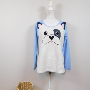 May include: A light blue and white long-sleeved shirt featuring a dog face design. The dog's face is white with black and pink accents. The shirt is displayed on a white mannequin.