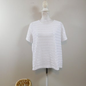 May include: White short-sleeved top with a textured, sequined design. The round-neck shirt is displayed on a mannequin. A woven basket with a rope handle sits in the foreground.