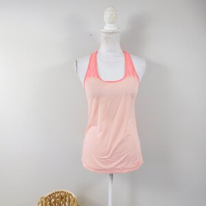 May include: A peach-colored tank top with a racerback design and a pink mesh panel on the back. The tank top is on a white mannequin.