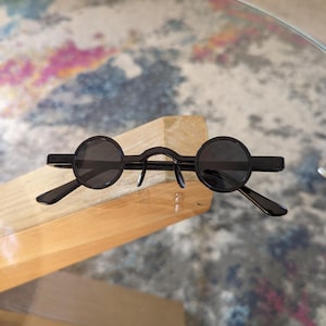 Peut inclure: Lunettes de soleil rondes noires avec une fine monture en métal. Les lunettes sont pliées en deux et posées sur une surface en bois.