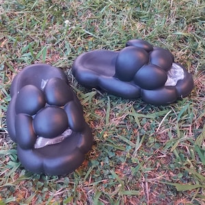 May include: Two black, cloud-shaped sandals rest on a bed of green grass. Each sandal features a series of rounded, bubble-like protrusions. The sandals appear to be made of a soft, rubbery material. One sandal has a piece of clear plastic inside.