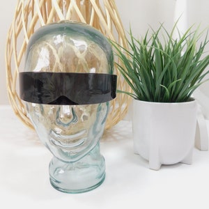 May include: A glass head mannequin with a black visor. The mannequin is sitting on a white surface. There is a wicker basket and a potted plant in the background.