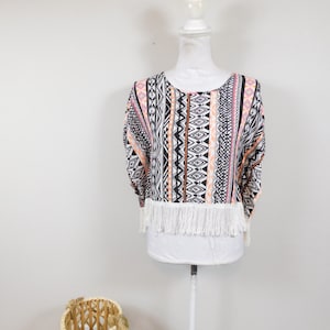 May include: A patterned top with a white fringe hem is displayed on a mannequin. The top features a mix of geometric patterns in black, white, pink, and orange. The mannequin is on a white stand, and a woven basket is in the foreground.
