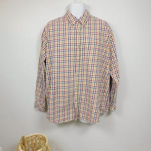 May include: A long-sleeved button-down shirt in a classic plaid pattern. The shirt features a button-down collar and a chest pocket. The plaid design incorporates red, yellow, and navy blue squares on a white background. The shirt is displayed on a white mannequin.