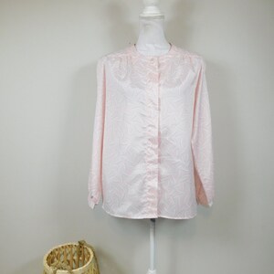 May include: A light pink long-sleeve blouse with a subtle, repeating pattern of gray lines. The blouse has a mandarin collar and button-down front. It is displayed on a white mannequin. A woven basket is in the foreground.