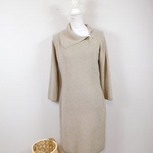 May include: A beige knitted dress with a cowl neck and a gold button detail. The dress is on a white mannequin.