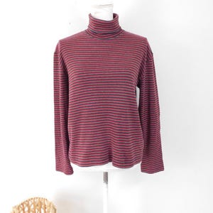 May include: A burgundy and black striped long-sleeved turtleneck shirt. The shirt is displayed on a mannequin against a white backdrop. The turtleneck has a thin blue stripe. The shirt's measurements are in centimetres.