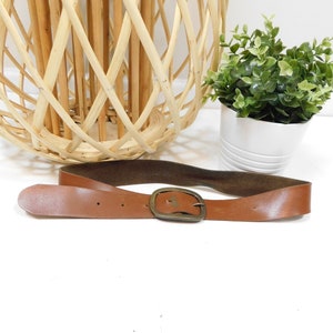 May include: A brown leather belt with a brass buckle. The belt is lying on a white surface.