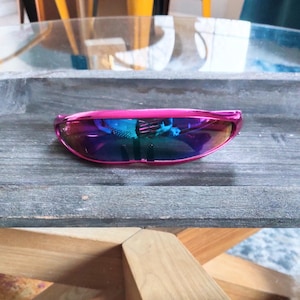 Könnte beinhalten: Eine futuristisch anmutende Sonnenbrille mit einem leuchtend pinkfarbenen Rahmen und schillernden Gläsern. Die Gläser reflektieren ein Farbspektrum und erzeugen einen auffälligen Look. Die Sonnenbrille liegt auf einer verwitterten Holzoberfläche.