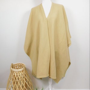 May include: A light tan poncho-style cape draped over a white mannequin. The cape has a V-neck and a flowing design. A woven basket and a small potted plant are in the foreground.