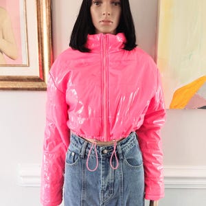 May include: A vibrant pink, cropped puffer jacket with a high collar and a front zip. The jacket features long sleeves and a drawstring waist. It is styled with blue denim jeans.