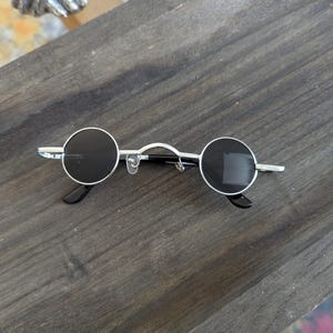 May include: Round sunglasses with silver frames and black lenses. The sunglasses have a simple, minimalist design.