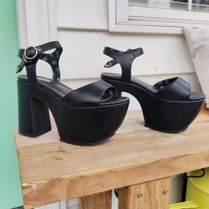 May include: A pair of black platform sandals with thick block heels. The sandals have an ankle strap with a silver buckle and a toe strap. The platform soles are approximately 7.6 cm high. The sandals are displayed on a wooden surface.