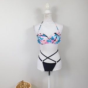 Könnte beinhalten: Ein zweiteiliger Badeanzug auf einer Schaufensterpuppe. Das Oberteil ist ein trägerloser Stil mit einem tropischen Blumendruck in Blau, Rosa und Schwarz. Die Unterseite ist einfarbig schwarz, mit gekreuzten Trägern. Die Schaufensterpuppe steht auf einem weißen Ständer.