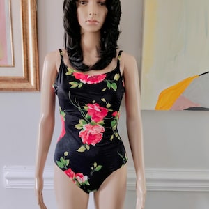 May include: A black one-piece swimsuit featuring a vibrant floral pattern with large pink roses and green leaves. The garment has thin shoulder straps, a scoop neckline, and a high-cut leg design, showcasing a bold botanical print on a dark base.