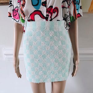 May include: A light blue and white checkered mini skirt. The skirt is made of a textured fabric and has a high waist. The model is wearing a white button-up shirt with a colorful abstract design.