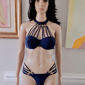May include: A dark blue two-piece bikini set featuring a high-neck halter top with multiple thin straps connected by gold-tone rings. The matching bikini bottoms include side cutouts with thin straps and gold-tone ring accents on both hips.