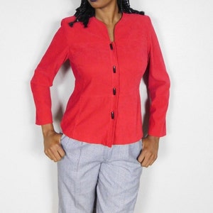 Könnte beinhalten: Ein leuchtend roter Blazer mit Stehkragen und langen Ärmeln. Der Blazer hat schwarze Knöpfe auf der Vorderseite. Die Person trägt eine blau-weiß karierte Hose.
