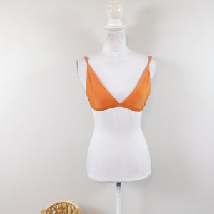 Puede incluir: Top de bikini naranja triangular exhibido en un maniquí blanco. El top tiene tirantes finos y un diseño sencillo. Una cesta de mimbre está en primer plano, con una pared blanca lisa en el fondo.