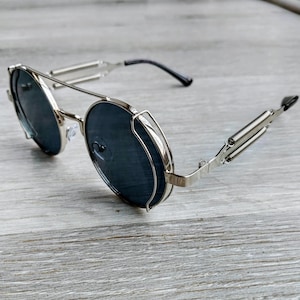 May include: Silver-framed sunglasses with dark lenses. The glasses have a unique steampunk design, featuring spring-loaded temples and a round frame. The lenses are a dark gray color, and the overall style is vintage-inspired.