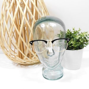 May include: Black and gold rimmed glasses on a glass mannequin head. The glasses have a browline style with a black top and gold bottom.