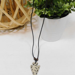 May include: A cream-colored arrowhead pendant necklace with a black cord. The pendant has dark brown etched lines and two small wooden beads. The necklace is displayed against a white background with a green plant in a white pot.