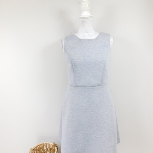 Puede incluir: Un vestido sin mangas, gris claro, con falda acampanada se exhibe en un maniquí blanco. El vestido tiene un corpiño ajustado y un escote redondo. Una cesta de mimbre está en el suelo en primer plano.