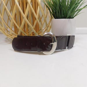 Può includere: Una cintura marrone scuro lucida con una grande fibbia argentata. La cintura ha un design classico e sembra essere realizzata in pelle. La fibbia è una caratteristica importante, che aggiunge un tocco di eleganza all'accessorio. Adatta a vari outfit.