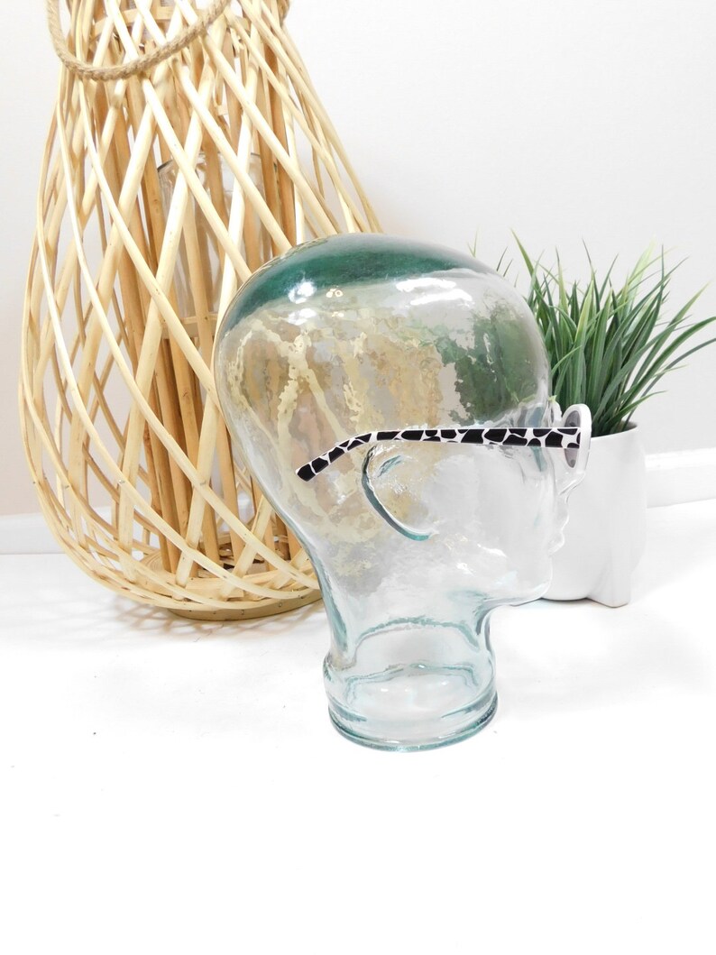 May include: A clear glass mannequin head with a black and white patterned eyeglass frame resting on the nose. The mannequin head is sitting on a white surface.
