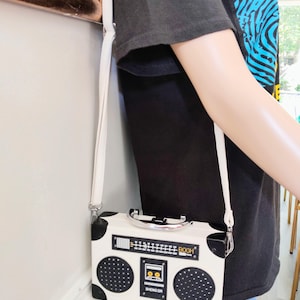 May include: A white and black crossbody bag shaped like a vintage boombox. The bag has a silver handle, a white strap, and the text "BOOM" and "SHENG DA" on the front. The bag is being worn over the shoulder.
