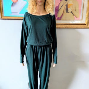 May include: A dark green long-sleeved jumpsuit with a relaxed fit and a wide neckline. The jumpsuit has a drawstring waist and cuffed legs.