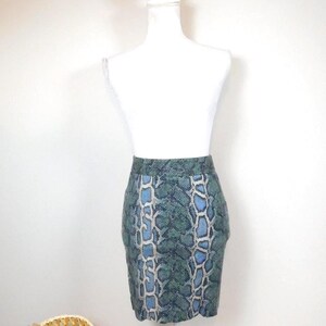 Puede incluir: Minifalda con estampado de piel de serpiente en tonos verde azulado, azul y beige. La falda se exhibe en un maniquí blanco. Una cesta de mimbre se encuentra en la esquina inferior izquierda. La falda mide unos 40 cm.