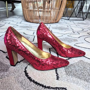 May include: A pair of red sequined high heels with a block heel. The shoes have a pointed toe and are made of a sparkly material.