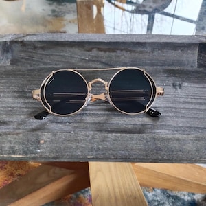 May include: Round sunglasses featuring dark lenses encased in a gold-toned metal frame with a double bridge design. The circular lenses are surrounded by a thin wire rim, and the temples are connected to the frame with visible metal hinges.