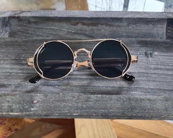 Peut inclure: Lunettes de soleil rondes dotées de verres sombres encadrés par une monture en métal doré avec un double pont. Les verres circulaires sont entourés d'un fin cerclage métallique et les branches sont fixées à la monture par des charnières visibles.