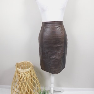 Puede incluir: Una falda de cuero marrón con silueta recta y superficie ligeramente texturizada. La falda se muestra en un maniquí blanco.