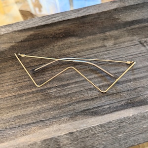 Puede incluir: Un par de gafas doradas con un diseño de marco angular único. Las gafas están dobladas y descansan sobre una superficie de madera.