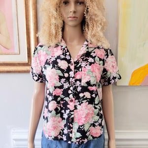 Puede incluir: Camisa negra con estampado floral rosa y blanco. La camisa de manga corta tiene un cuello clásico y un bolsillo en el pecho. El estampado floral presenta grandes flores rosas con hojas verdes sobre un fondo negro. La camisa se combina con vaqueros azules.