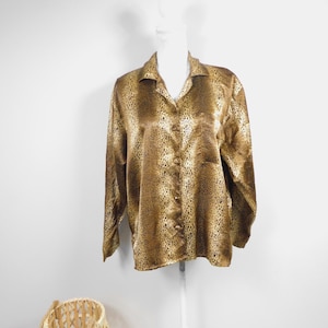 Puede incluir: Camisa abotonada de manga larga con estampado de leopardo en tonos marrones y beige. La camisa se exhibe en un maniquí blanco. Una cesta de mimbre se encuentra en la esquina inferior izquierda.