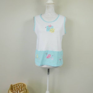 May include: White sleeveless top with a blue and white striped trim. The top has a small embroidered design of a gift box with flowers on the front and a blue and white striped band with embroidered flowers at the bottom.