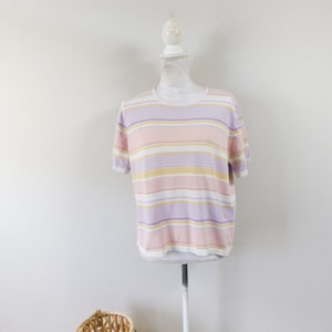 May include: A short-sleeved, pastel-striped shirt in shades of pink, lavender, and yellow. The shirt is displayed on a white mannequin. The shirt has a round neckline and a relaxed fit. A woven basket is in the foreground.