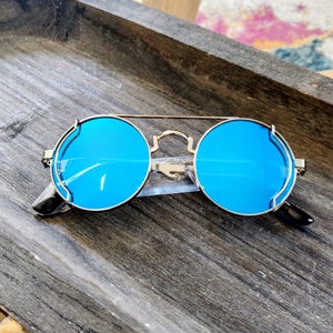 May include: Round sunglasses with bright blue lenses and a gold-colored metal frame. The sunglasses have a double bridge and black earpieces. The lenses are reflective and the sunglasses are resting on a wooden surface.
