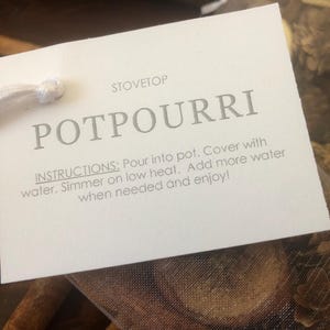 Może przedstawiać: Biała etykieta z szarym napisem "POTPOURRI" oraz instrukcją użytkowania. Etykieta jest przymocowana do siatkowego worka wypełnionego suszonymi roślinami i laskami cynamonu.