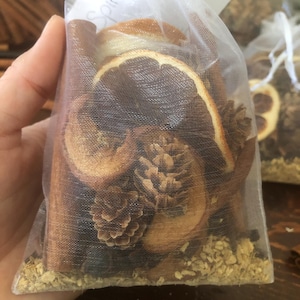 Può includere: Un sacchetto di rete bianca pieno di fette di arancia essiccate, bastoncini di cannella, pigne e altri ingredienti naturali. Il sacchetto è etichettato "Spice".