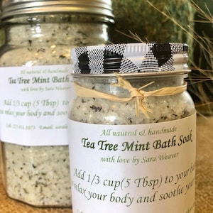 May include: Two glass jars filled with a white bath soak with green and brown herbs. The jars have black and white checkered lids and labels that read "All natural & handmade Tea Tree Mint Bath Soak with love by Sara Weaver". The labels also include instructions to add 1/3 cup (5 Tbsp) to your bath to relax your body and soothe your skin.