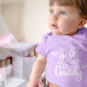 Może przedstawiać: Dziecko w liliowym body z napisem "Just a girl in love with her Daddy" w białym kolorze.