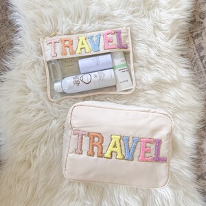 May include: Two beige travel pouches with the word "TRAVEL" embroidered in colorful letters. The top pouch is clear and contains several toiletries.