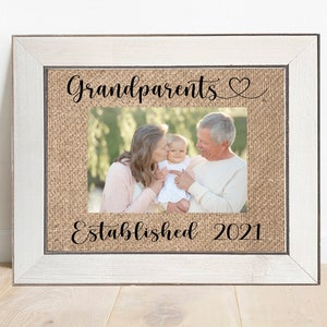 May include: A white picture frame with a burlap mat and a photo of grandparents holding a baby. The text "Grandparents" and a heart symbol are at the top of the mat. The text "Established 2021" is at the bottom of the mat.